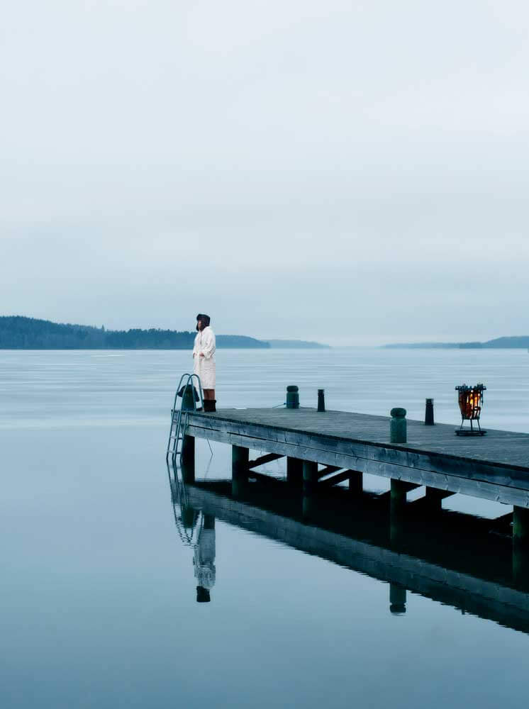 Kvinna i vit morgonrock längst ut på en brygga i blå isig vintrig miljö med eld i en eldkorg