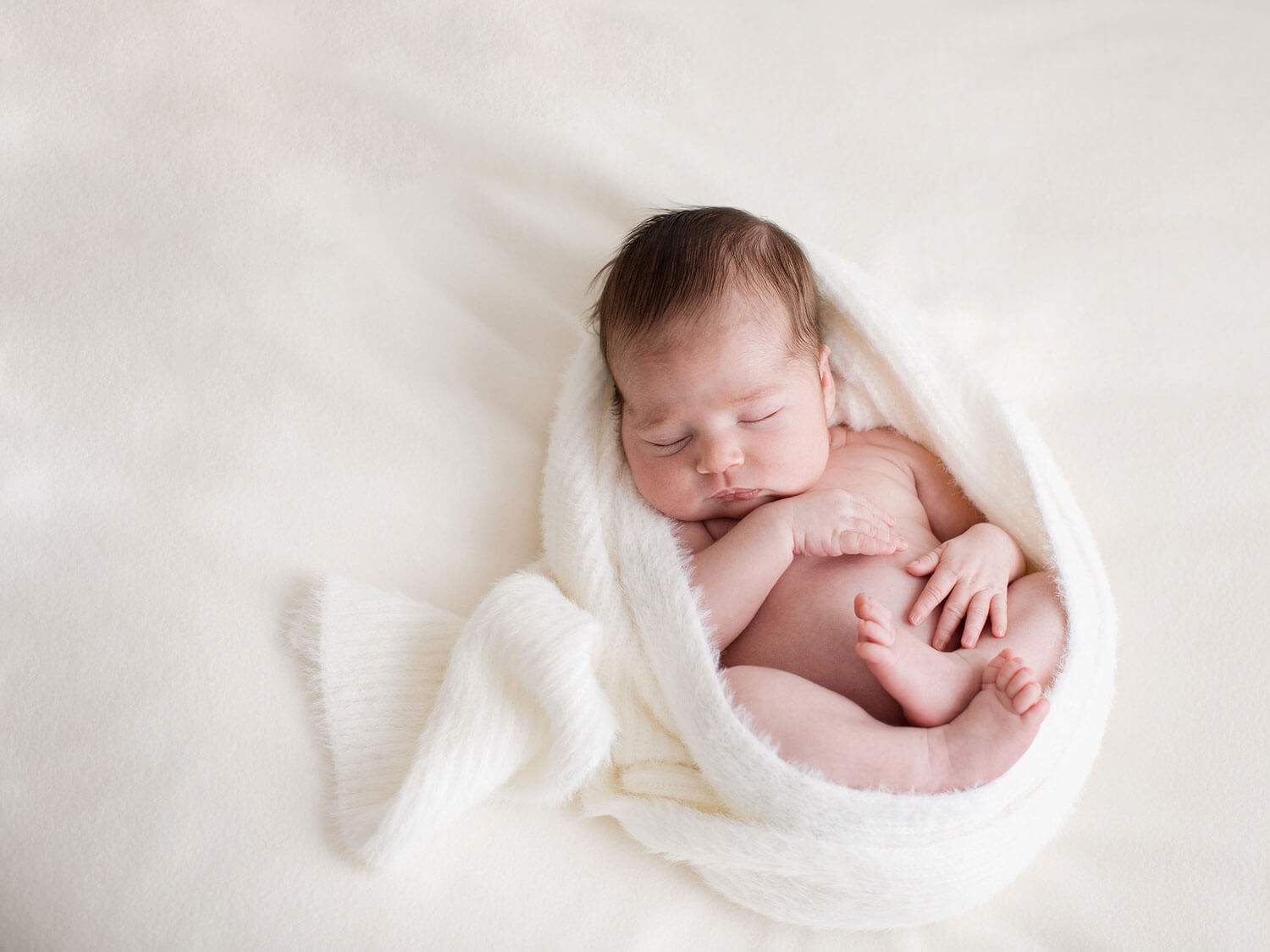 nyfödd bebis sovande på en ljus kudde med bar kropp inlindad i en vit fluffig sjal